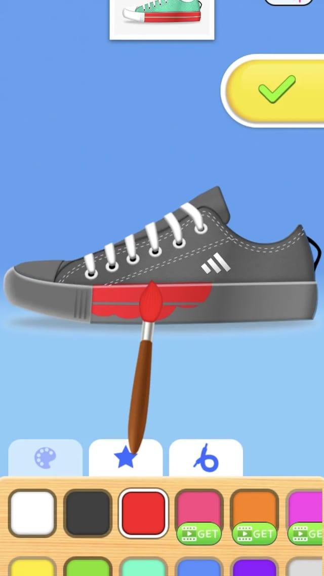 Shoes Maker design screen showing a grey canvas sneaker being painted with red brush strokes, a stripe decal applied on its side, a reference photo of the target sneaker above, a checkmark button at the right and a color swatch palette below